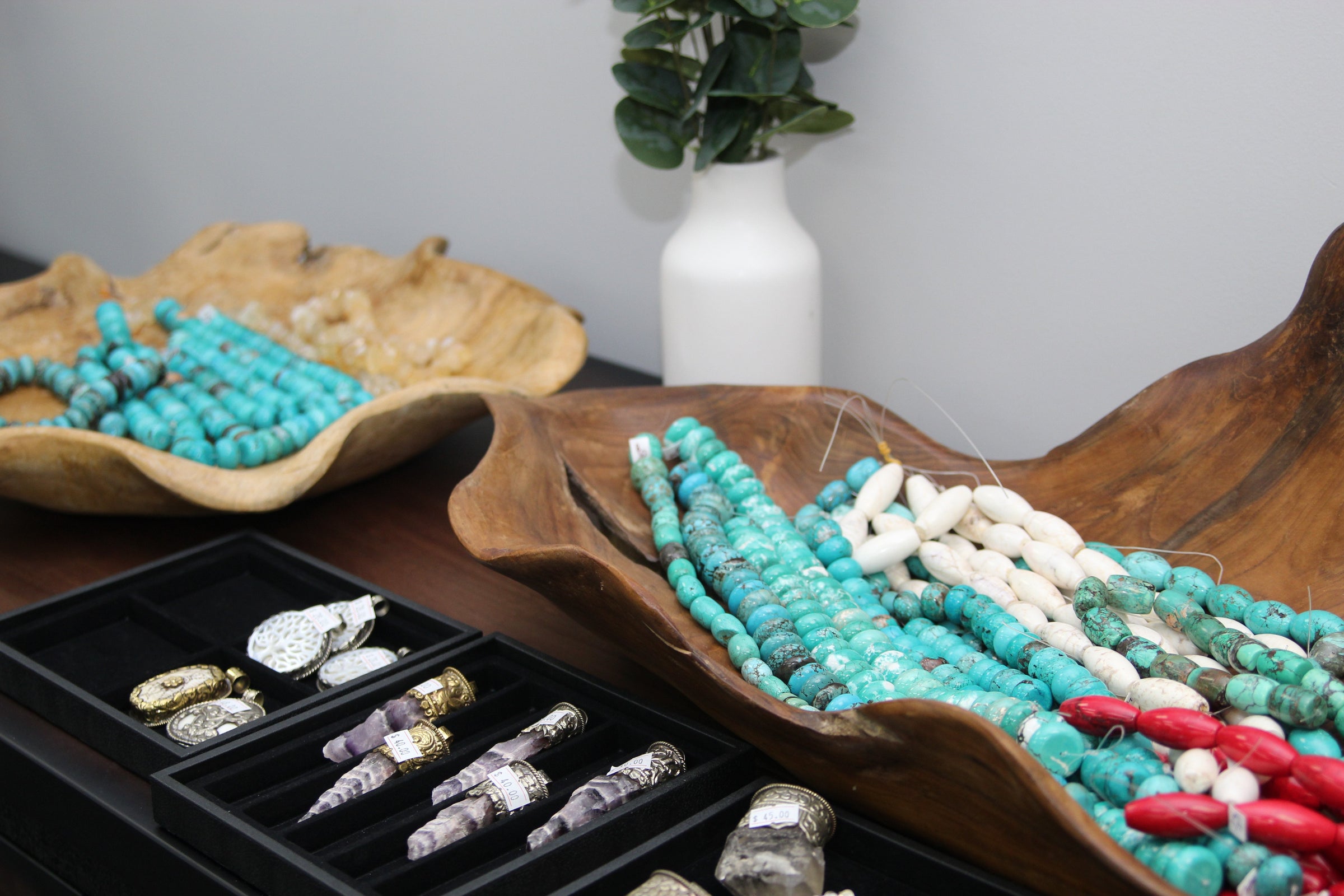 Turquoise beads in a wooden bowl sit on a table with trays of amethyst and bone pendants available at the Crafting District. The Crafting district is where makers go to find beautiful gemstones, books, jewelry making and craft supplies.  
